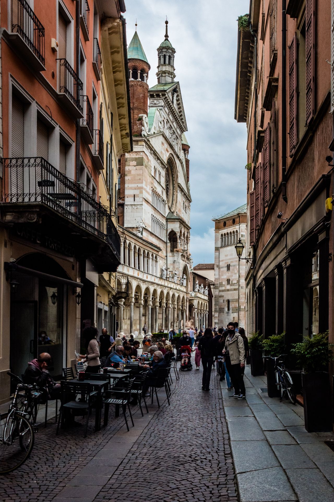 Narrow cobblestone street with outdoor cafe tables and the cathedral campanile visible at the end, Cremona, Lombardy, Italy