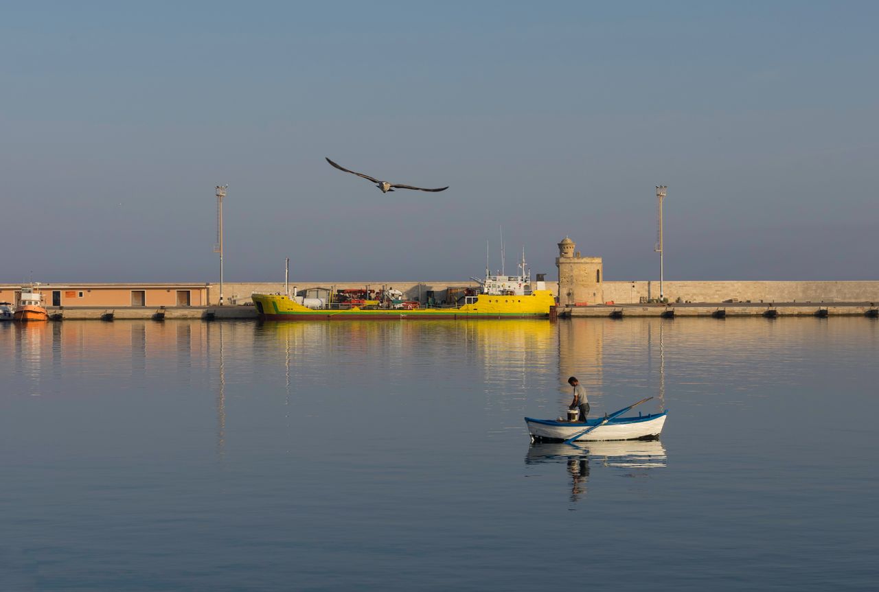 Small fishing boat in the calm blue waters of Gallipoli harbour with the historic old town walls behind, Puglia, Italy
