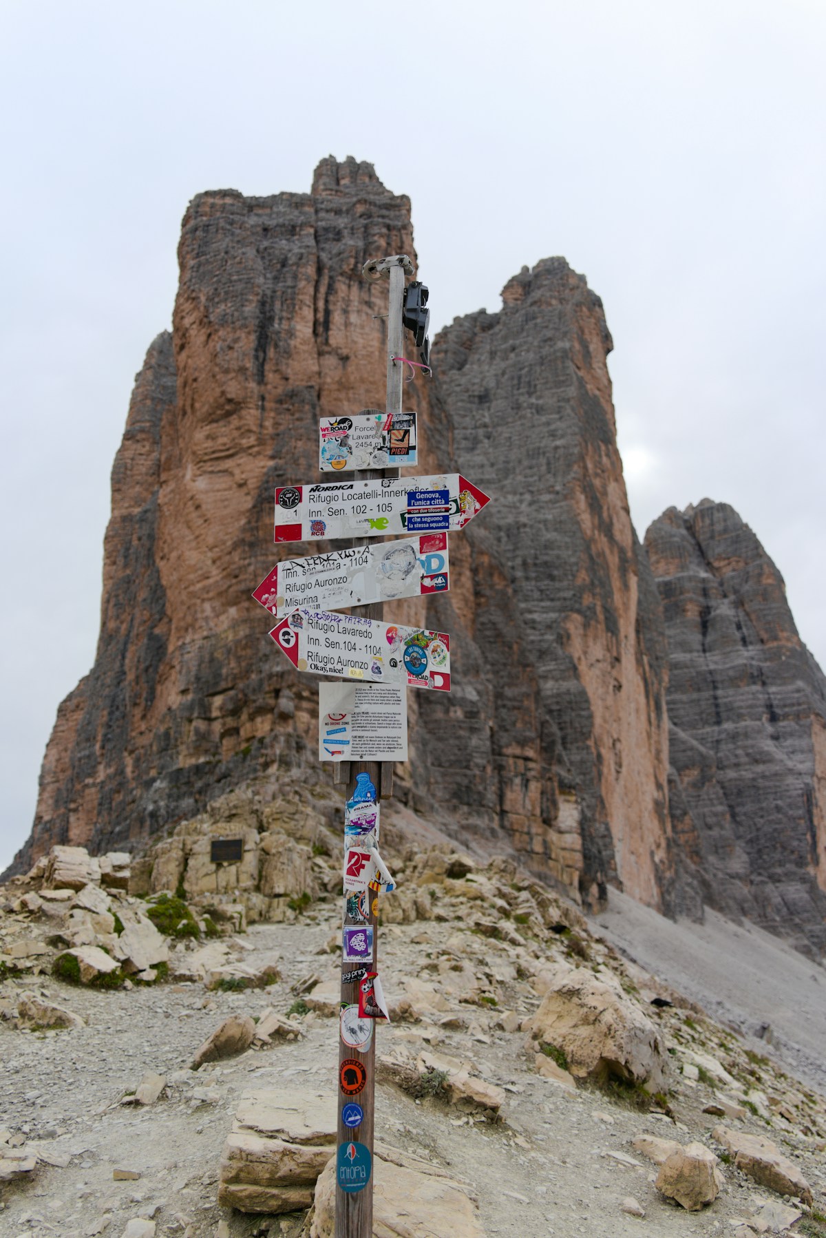 Wooden trail signpost covered in route markers in front of the three rocky peaks of Tre Cime di Lavaredo, Dolomites, Italy