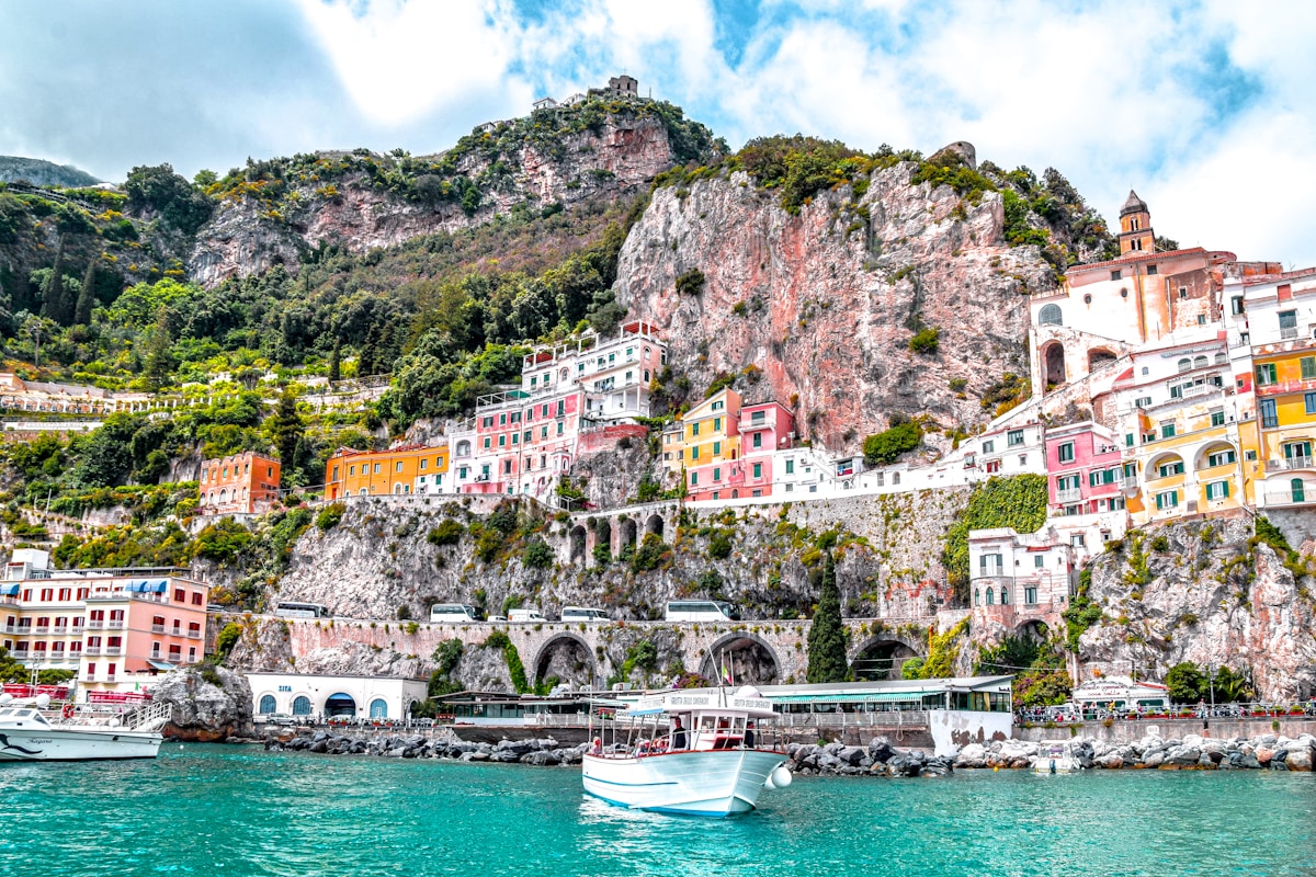 White boat on turquoise water below colourful cliffside buildings of the Amalfi Coast, Campania, Italy