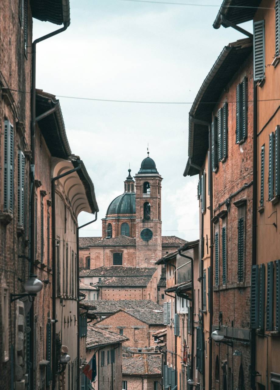 Narrow alley of terracotta-coloured buildings with the Urbino cathedral dome visible at the end, Le Marche, Italy