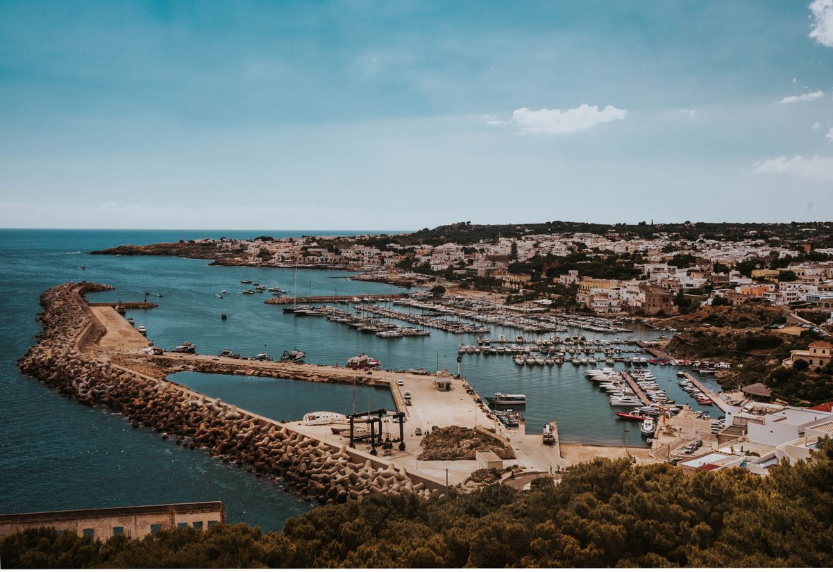 Aerial view of a Puglia coastal town with a curved harbour, breakwater, and boats moored in clear Ionian water, southern Italy