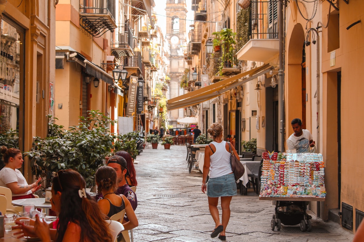 People walking past outdoor cafes and historic buildings on a busy Palermo street, Sicily, Italy