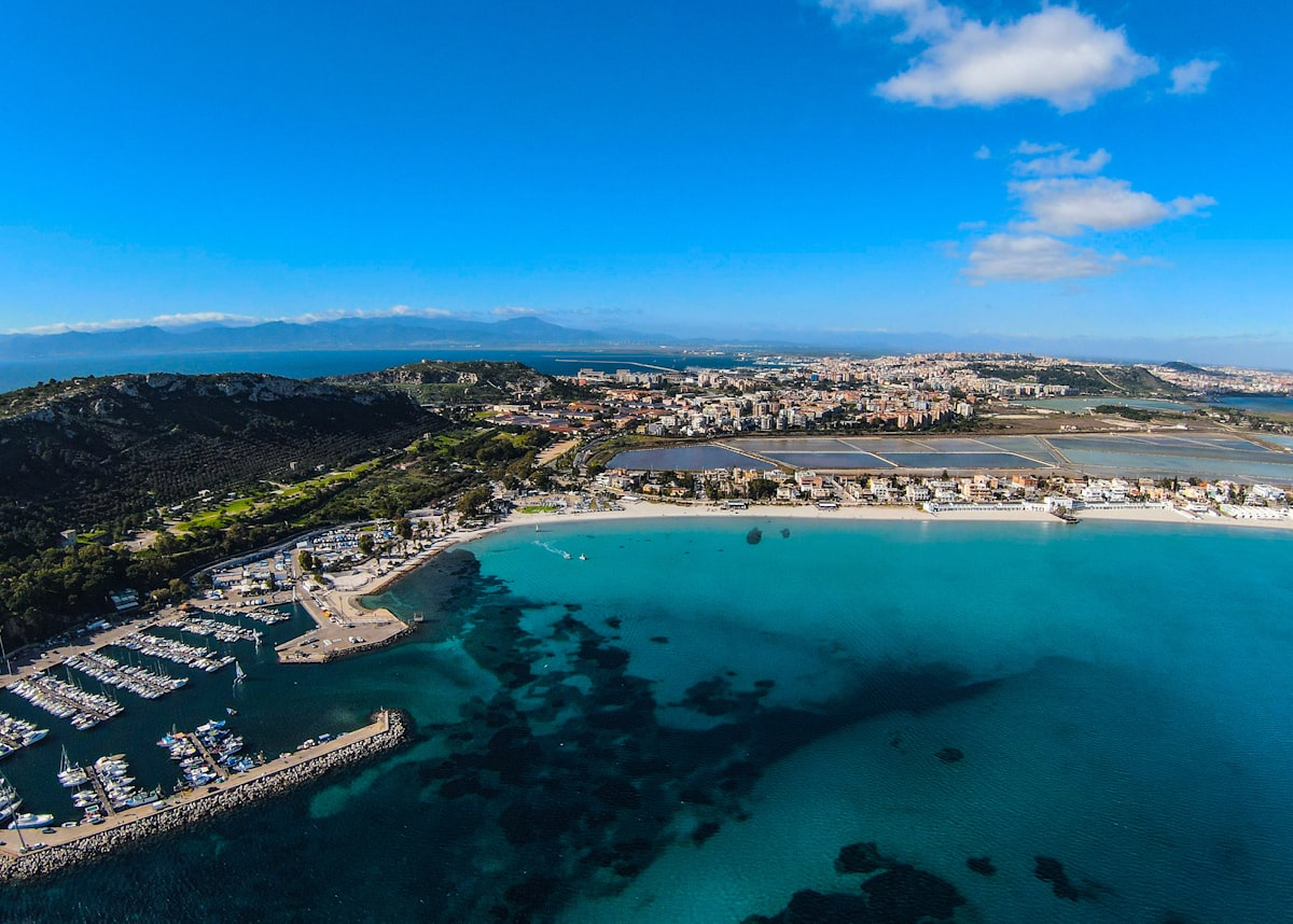 Aerial view of a Sardinian marina and beach with turquoise water, white sand, and a coastal town stretching to the horizon
